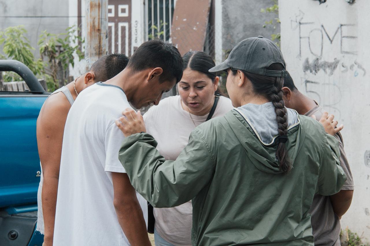 Volunteers and neighbors standing in a circle with hands on shoulders, praying together on a street