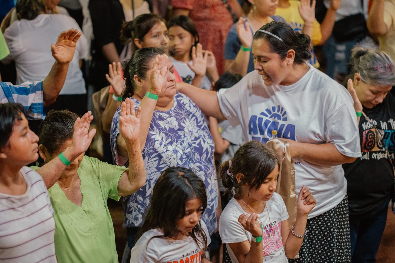A group of women, children, and neighbors with raised hands receiving prayer during an outreach gathering